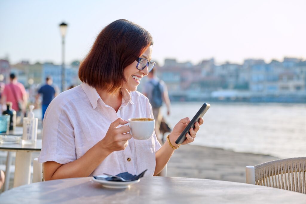 Learn Greek Hub middle aged woman resting in outdoor cafe with a cup of coffee using a smartphone